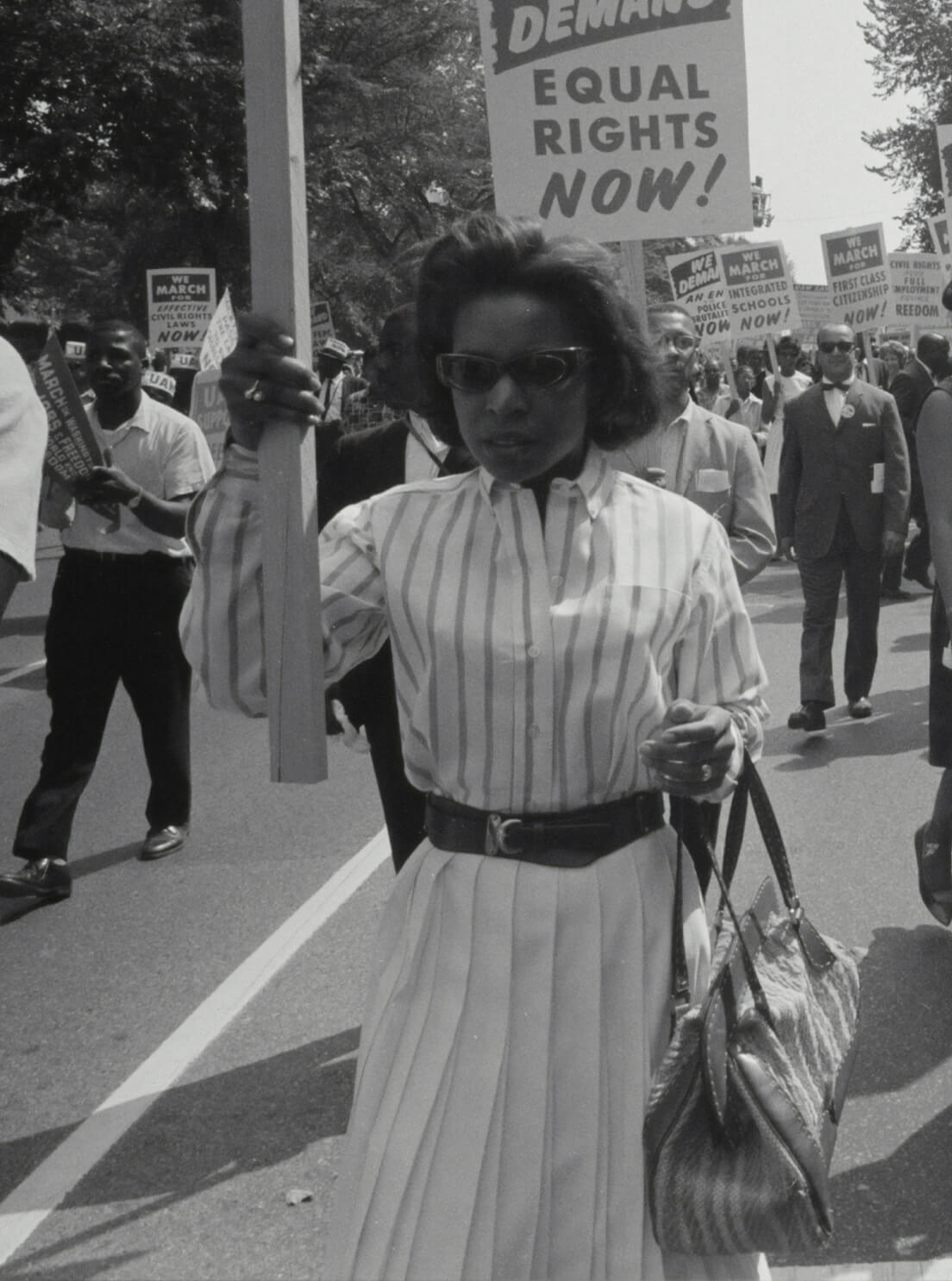 A black-and-white photo from the 1963 March on Washington featuring a woman holding a large sign that reads, "WE DEMAND EQUAL RIGHTS NOW!"