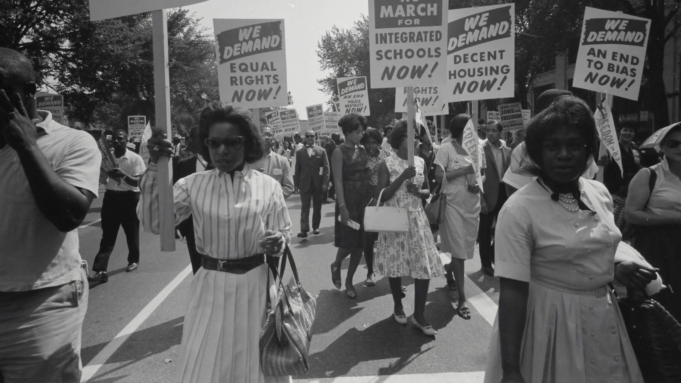 A black-and-white photo from the 1963 March on Washington featuring a woman holding a large sign that reads, "WE DEMAND EQUAL RIGHTS NOW!"